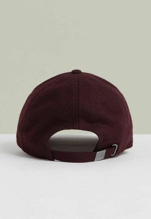 Bordo krāsas regulējamā beisbola cepure redzama no aizmugures ar metāla sprādzi un auduma siksniņu uz baltas virsmas un gaiši pelēka fona.