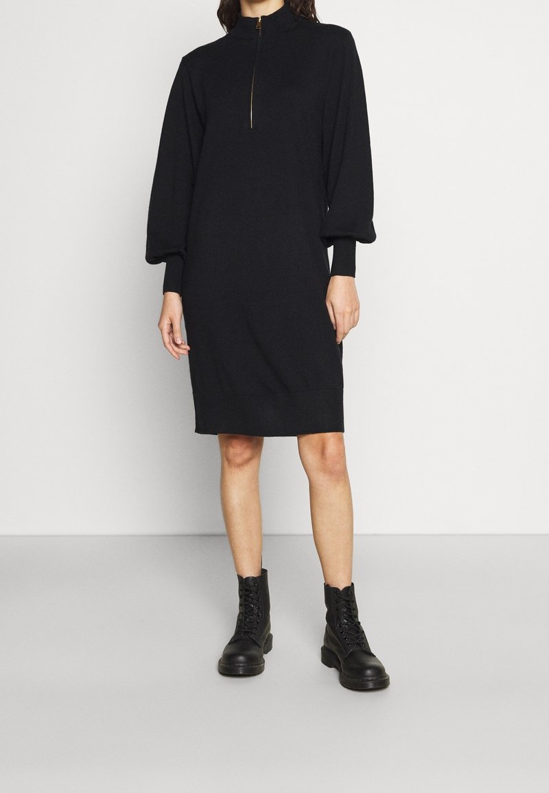 Robe noire avec un col montant et une demi-fermeture éclair, dotée de longues manches bouffantes, de poignets côtelés et d'une silhouette droite. Portée avec des bottes de combat noires.