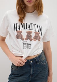 Maglietta bianca in cotone con una grafica che presenta tre orsetti di peluche marroni, con la scritta "MANHATTAN" e "NEW YORK 1994" stampata in grassetto nero.