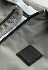Siva jakna z gladkim, odbojnim materialom, zadrgo in črnim gumijastim znakom z odtisnjenim besedilom "BAKER BY TED BAKER."