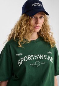 Zielona bawełniana koszulka z napisem "SPORTSWEAR" w białym, pogrubionym tekście oraz logo "hummel". Granatowy kapelusz z napisem "SPORTSWEAR SINCE 1923" w białym kolorze.