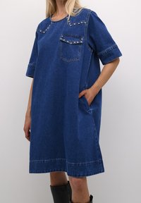 Jeansklänning i medelblått med korta ärmar, rundad halsringning, framfickor och dekorativa nitar på axlarna och fickan.