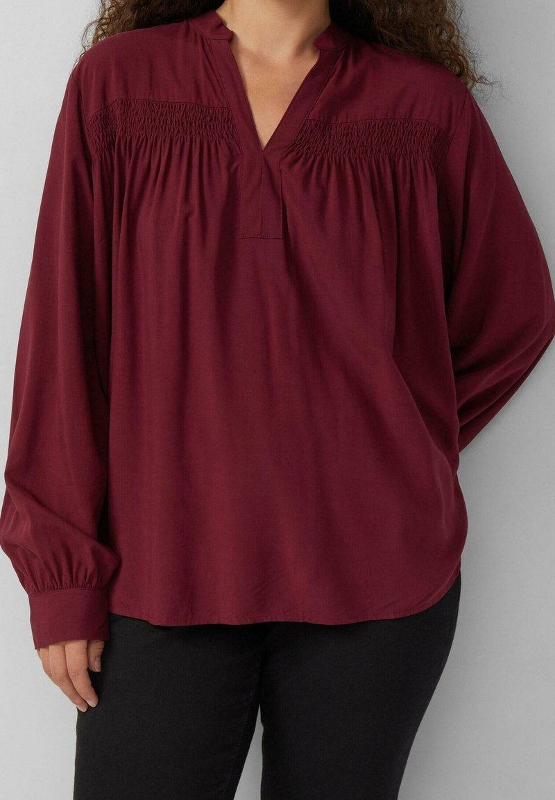 Femme portant un chemisier amplement ajusté à manches longues de couleur bordeaux avec des détails froncés sur la poitrine et un pantalon noir, sur un fond uni.