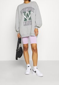 Šedá oversized mikina s potiskem "WESTFORT LEAGUE", v kombinaci s lila cyklistickými šortkami a bílými teniskami Nike. Černá taška se strapcem.