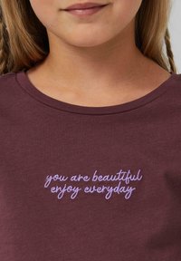 Burgunderfarbene Baumwoll-T-Shirt mit lila gesticktem Text: "Du bist schön, genieße jeden Tag." Klassischer Rundhalsausschnitt und weicher Stoff.