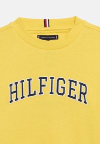 Κίτρινο φούτερ με στρογγυλή λαιμόκοψη και τη λέξη «HILFIGER»  κεντημένη με μπλε σκούρα γράμματα πλακώματος στο στήθος, καθώς και ετικέτα με το εμπορικό σήμα στο λαιμό.