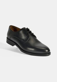 Chaussure de ville en cuir noir avec un bout arrondi, à lacets et un léger talon, présentant une texture lisse et peu d'ornementations.