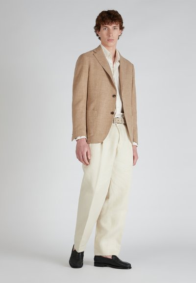 Lichtbruin linnen blazer met twee knopen, gecombineerd met crèmekleurige broeken en zwarte loafers. Geweven stof met subtiele patroondetails.
