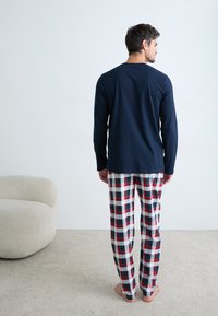 Man staat blote voeten op een vloerbedekking in een marineblauw overhemd met lange mouwen en rood, wit en groen geruite pyjamabroek naast een beige stoel.