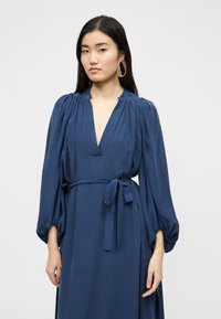 Γυναίκα με μακριά σκούρα μαλλιά φοράει ένα βαθύ μπλε φόρεμα με ντεκολτέ σε σχήμα V, μακριά φουσκωτά μανίκια δεμένα στη μέση και μεγάλα χρυσά οβάλ σκουλαρίκια.