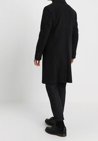 Cappotto nero con un design su misura, realizzato in un materiale testurizzato. Presenta un taglio lungo e polsini con bottoni. Indossato sopra pantaloni e scarpe scure.
