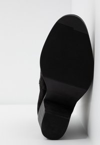 Semelle de botte noire avec bande de roulement en caoutchouc texturé, présentée à la verticale sur un fond blanc en angle.