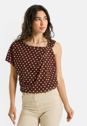 Femme aux longs cheveux foncés portant un top marron à pois à une épaule et un pantalon beige, regardant légèrement sur le côté.