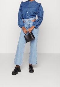 Jeansskjorta med volanger vid axlarna, knäppt framtill, kombinerad med ljusblå högmidjade jeans och svarta chunky klackskor, håller en liten svart väska.