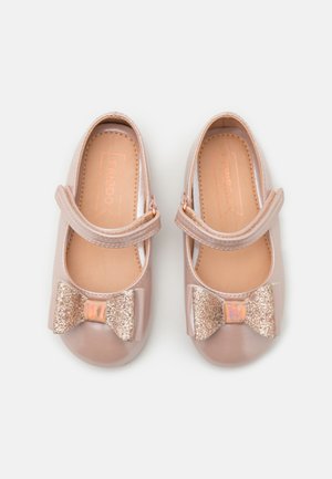 Par blanke beige småbørns sko med glitrende sløjfer og justerbare stropper på en ensartet hvid baggrund.