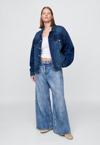 Jeans jakna in široke kavbojke. Temno modra tkanina z sproščenim krojem, sprednjimi žepi in gumbnim zapiranjem. Svetlo modre oprane kavbojke z ravnim krojem.