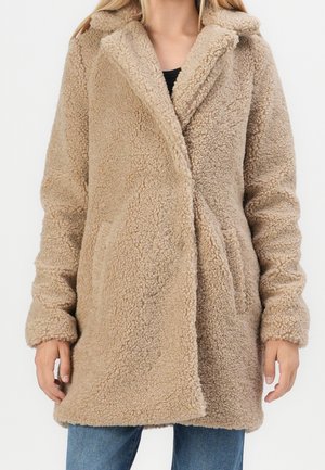 Femme portant un manteau teddy beige texturé à manches longues avec des poches latérales, par-dessus un jean bleu et un haut noir.