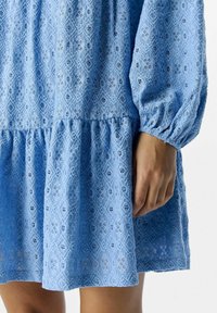 Helles blaues Kleid aus strukturiertem Stoff mit spitzenartigen Mustern, das über einen gerafften, gestuften Rock und lange puffärmel verfügt.