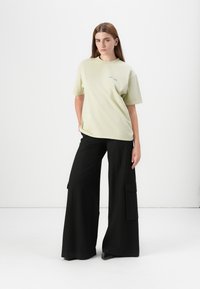 Model stands confidently, wearing a light green oversized t-shirt and black wide-leg cargo pants, set against a plain white background.