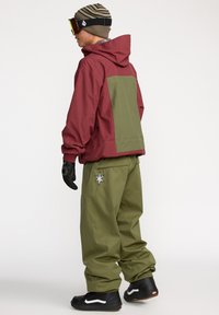 Rote und olivgrüne wasserdichte Jacke mit verstellbarem Kapuzen, kombiniert mit lockeren grünen Hosen mit einem Stern-Patch, schwarzen Handschuhen und Turnschuhen.