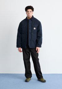 Marineblaue Arbeitsjacke mit vier Fronttaschen, einem Cordkragen und Metallverschlüssen, kombiniert mit schwarzen Hosen und grünen Schuhen.