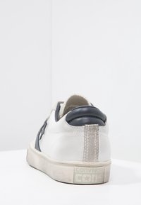 Baskets en cuir blanc avec accents bleu marine, talon gris texturé, et semelle en caoutchouc. Le logo est affiché à l'arrière du talon en vert clair.