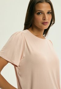 Różowa bluzka z krótkim rękawem wykonana z miękkiego materiału, z okrągłym dekoltem i marszczeniami na ramionach. Gładka, lekka tekstura.