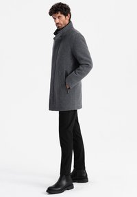 Abrigo de lana gris con un alto cuello, bolsillos laterales y una silueta recta. Combinado con pantalones negros ajustados y botines.