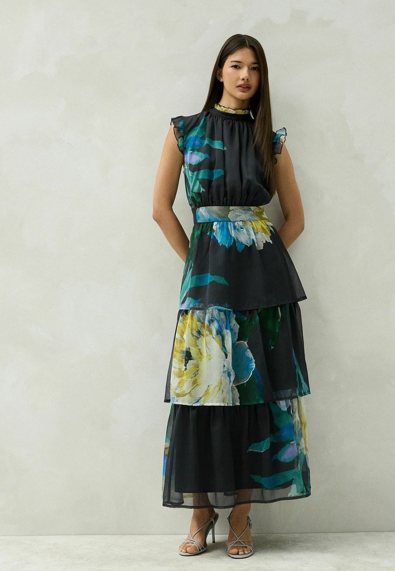 Woman with long dark hair wearing a sleeveless black tiered maxi dress with large blue, green, and yellow floral prints and silver heeled sandals.