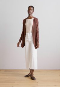 Brown long-sleeve cardigan over a light beige top, white wide-leg pants, and dark embellished flats on a wood floor with a neutral background.