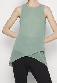 Helles grünes ärmelloses Oberteil aus fließendem, durchscheinendem Stoff, mit einem asymmetrischen Saum und einem runden Ausschnitt, kombiniert mit schwarzen Hosen.