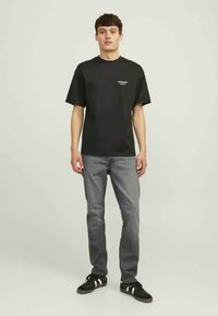 T-shirt noir à manches courtes avec un col rond, arborant un petit logo blanc sur la poitrine, associé à un jean gris et des baskets noires.