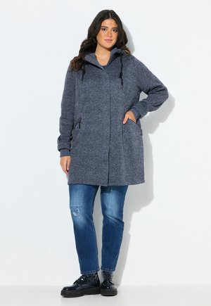 Kvinde iført en lang grå hættetrøje med lynlås og lommer, blå jeans og sorte støvler, stående med den ene hånd i lommen mod en hvid baggrund.
