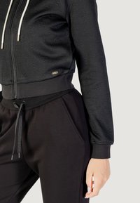 Felpa nera corta con tessuto testurizzato, chiusura con zip e cordini bianchi, abbinata a pantaloni jogger neri con tasche laterali e cordino.