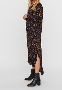 Mujer con un vestido largo negro y marrón con un estampado, que tiene una abertura lateral, llevando botas de cuero negras hasta el tobillo con tacones cuadrados, de pie de perfil.