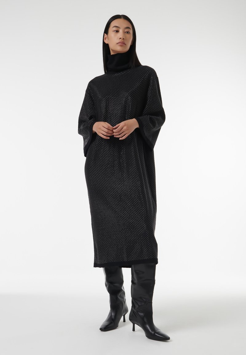 Vestido largo negro con cuello alto y un patrón geométrico texturizado. Mangas acampanadas y una silueta ajustada, combinado con botas negras hasta la rodilla.