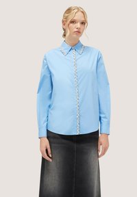 Cămașă albastră deschis cu mânecă lungă, cu guler ascuțit și plachetă cu nasturi ornamentată, asortată cu o fustă din denim neagră.