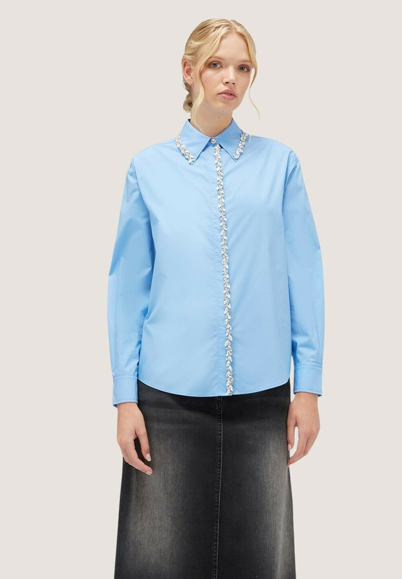 Camisa de manga larga azul claro con collar en punta y un tabique de botones decorado, combinado con una falda de mezclilla negra.