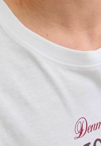 Primer plano de una camiseta blanca de cuello redondo usada por una persona, mostrando parte del cuello y texto rojo parcial en la camiseta.