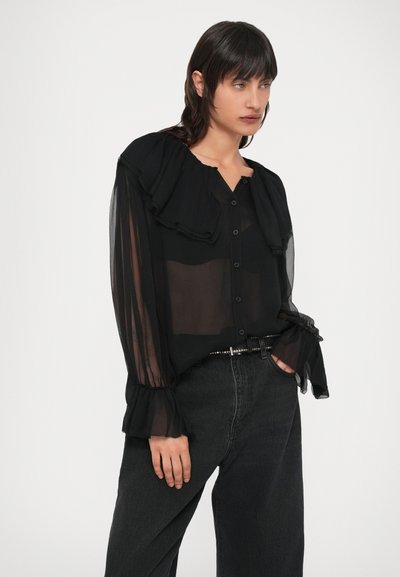 Blusa preta transparente com gola franzida e abotoamento frontal. Mangas compridas franzidas, combinada com jeans escuros de cintura alta e um cinto decorativo.