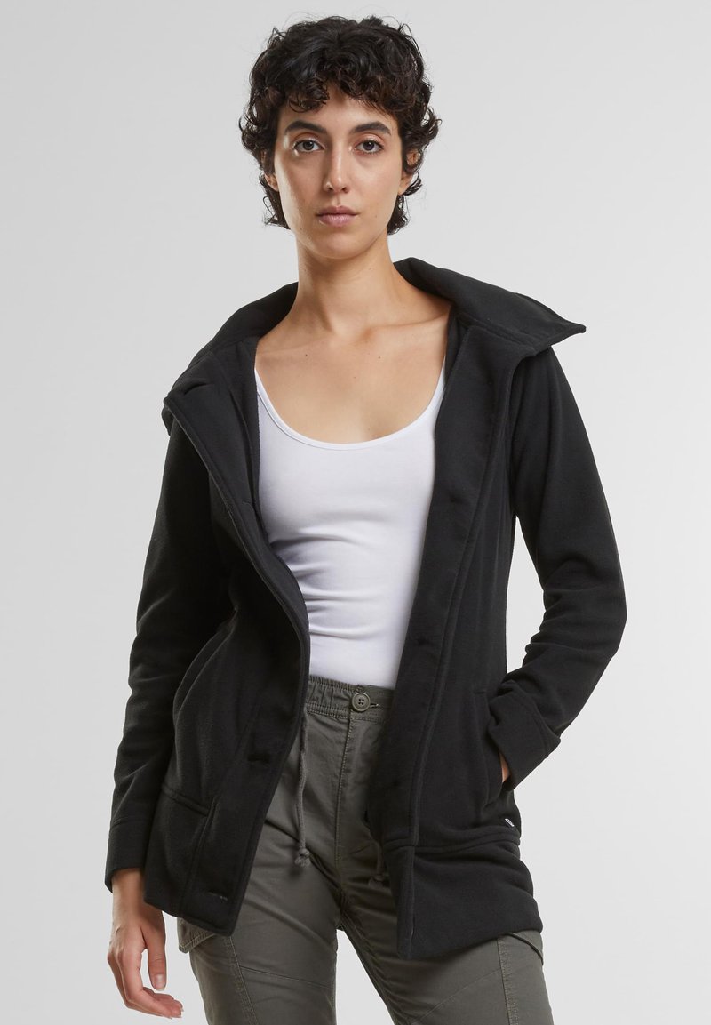 Črna flis jakna s kapuco, z zadrgo spredaj, dvema stranskima žepoma in mehko teksturo, nošena preko bele majice brez rokavov.