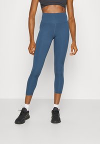 Högmidjade teal leggings i ett mjukt, stretchigt material med en åtsittande design, som når till mitten av vaden, tillsammans med svarta träningsskor.