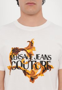Balta medvilnės marškinėliai su centriniu ornamentiniu auksiniu ir juodu spiralių grafiku, ant kurio įspaustas tekstas "VERSACE JEANS COUTURE".