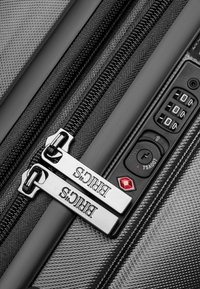 Maleta rígida negra con cremallera que presenta tiradores de metal plateado con marca, superficie texturizada, candado de combinación y emblema de seguridad aprobado por la TSA.