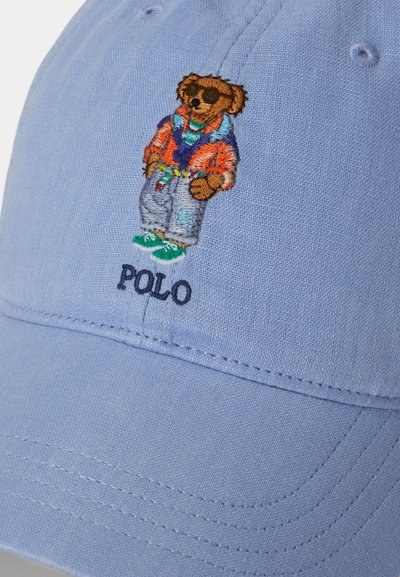 Ανοιχτό μπλε καπέλο με κεντημένη αρκούδα που φορά γυαλιά ηλίου, πορτοκαλί μπουφάν, τζιν, πράσινα αθλητικά παπούτσια και το κείμενο "POLO" κάτω από την αρκούδα.