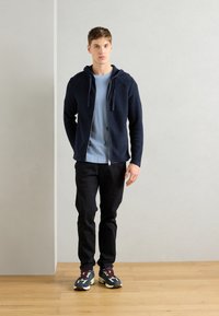 Felpa con zip blu navy e cordini su una maglietta azzurro chiaro, abbinata a pantaloni neri e sneakers multicolori su un pavimento in legno.