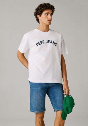 Jeune homme portant un t-shirt blanc "PEPE JEANS" et un short en jean, tenant une casquette de baseball verte dans sa main droite sur un fond uni.