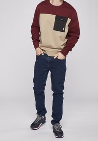Felpa beige e bordeaux con una tasca nera, abbinata a jeans blu scuri e sneakers nere. Texture liscia, design casual.