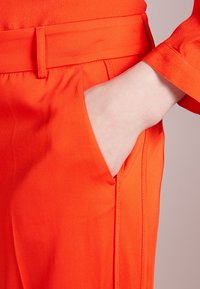 Pomarańczowe modne spodnie o gładkiej fakturze, z bocznymi kieszeniami i widocznymi szwami. Brak widocznego okucia lub wzorów.