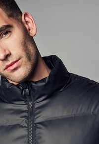 Chaqueta acolchada negra con un alto cuello y cierre frontal. La tela tiene una textura suave y brillante, con detalles de diseño minimalistas.